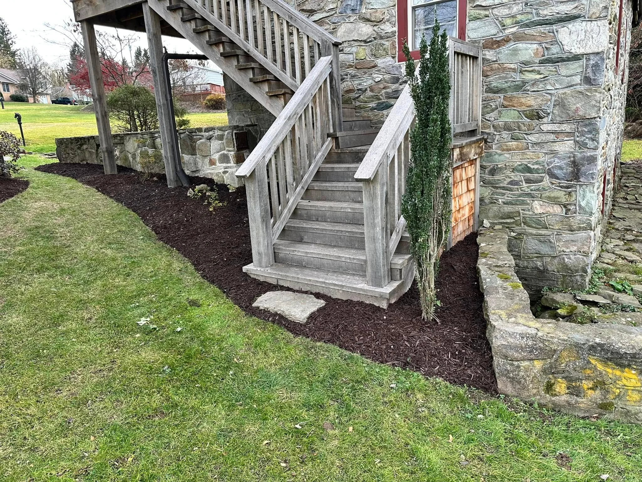 Dark mulch landscape beds around stone home foundation, wooden deck stairs, and lawn edge
