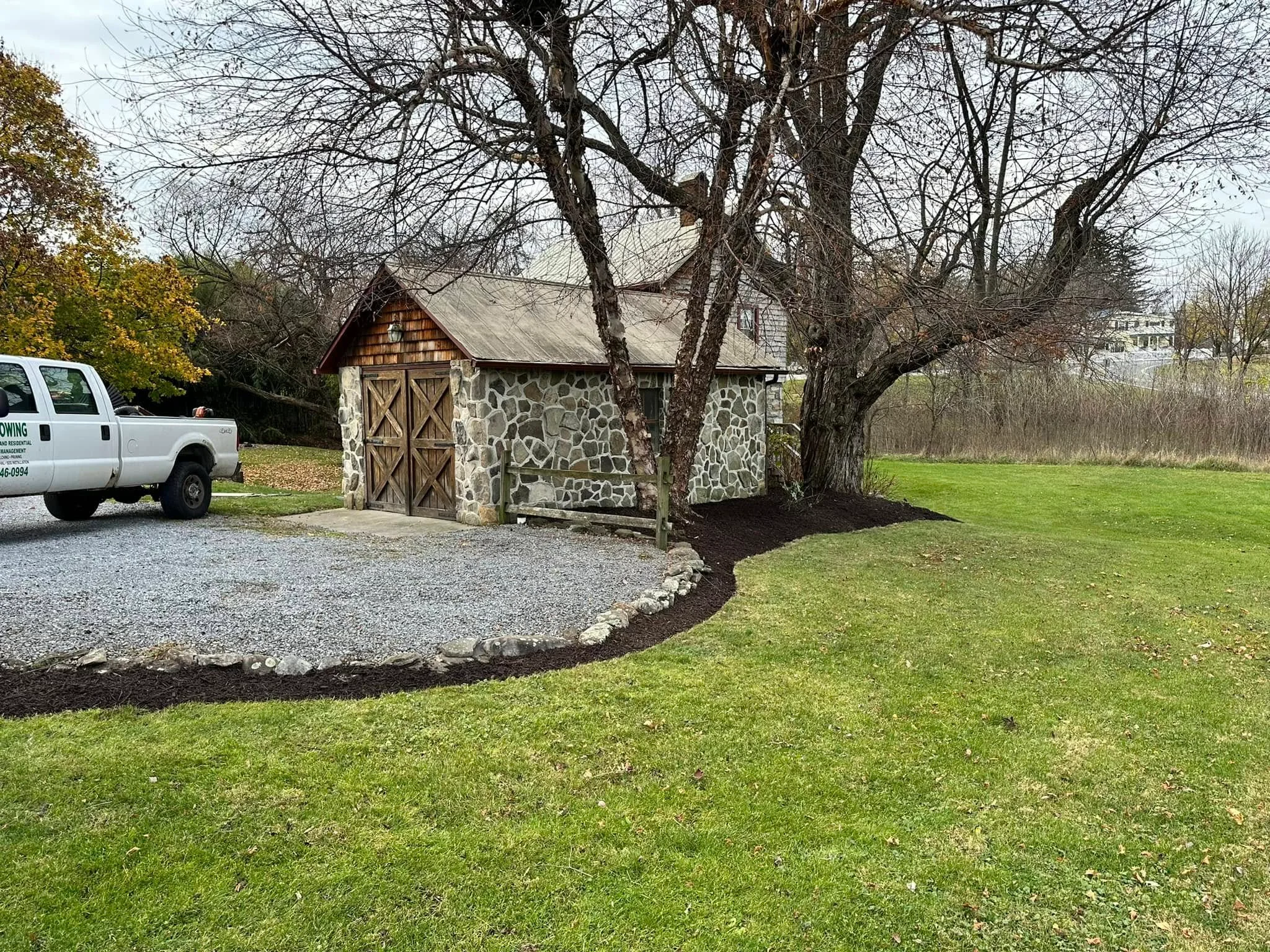 Gravel driveway with stone border edging, mulch beds, and lawn around stone cottage with mature trees