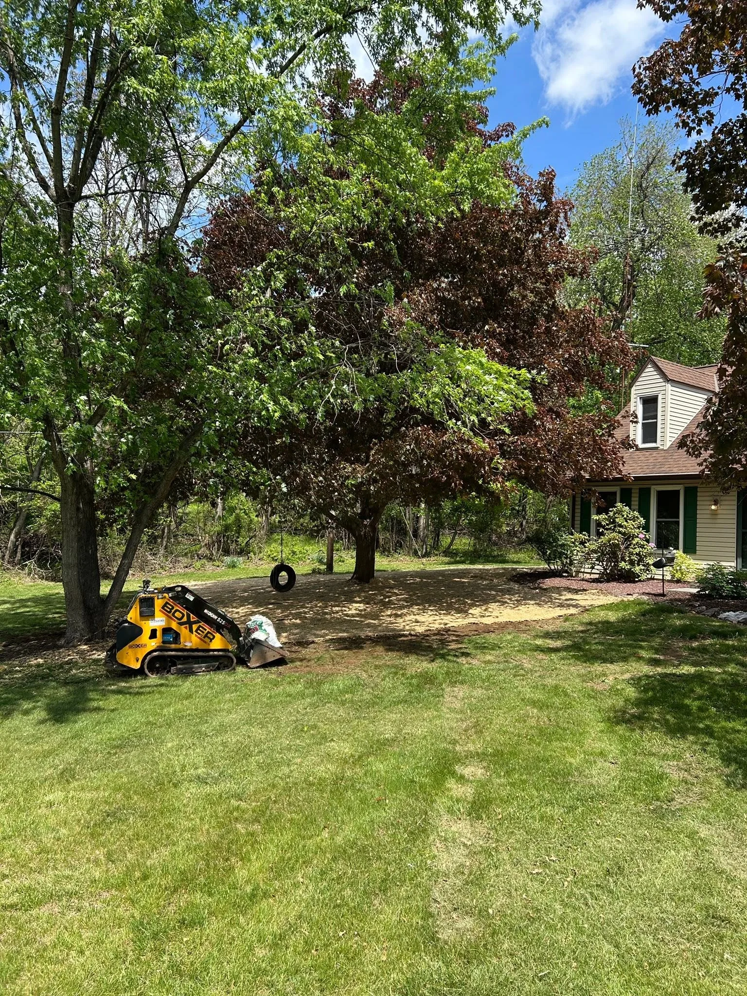Yellow compact track loader clearing residential yard with mature trees and house in background