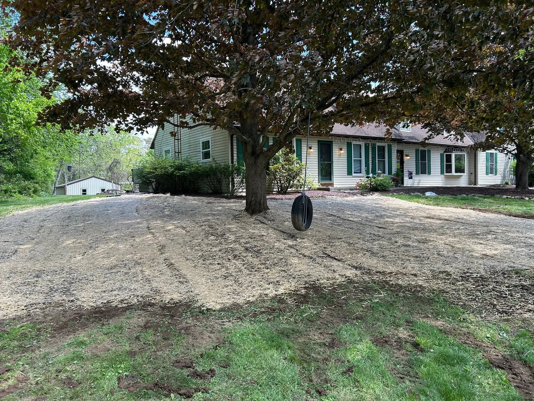 Graded residential yard with mature oak tree, prepared soil surface, and white siding home